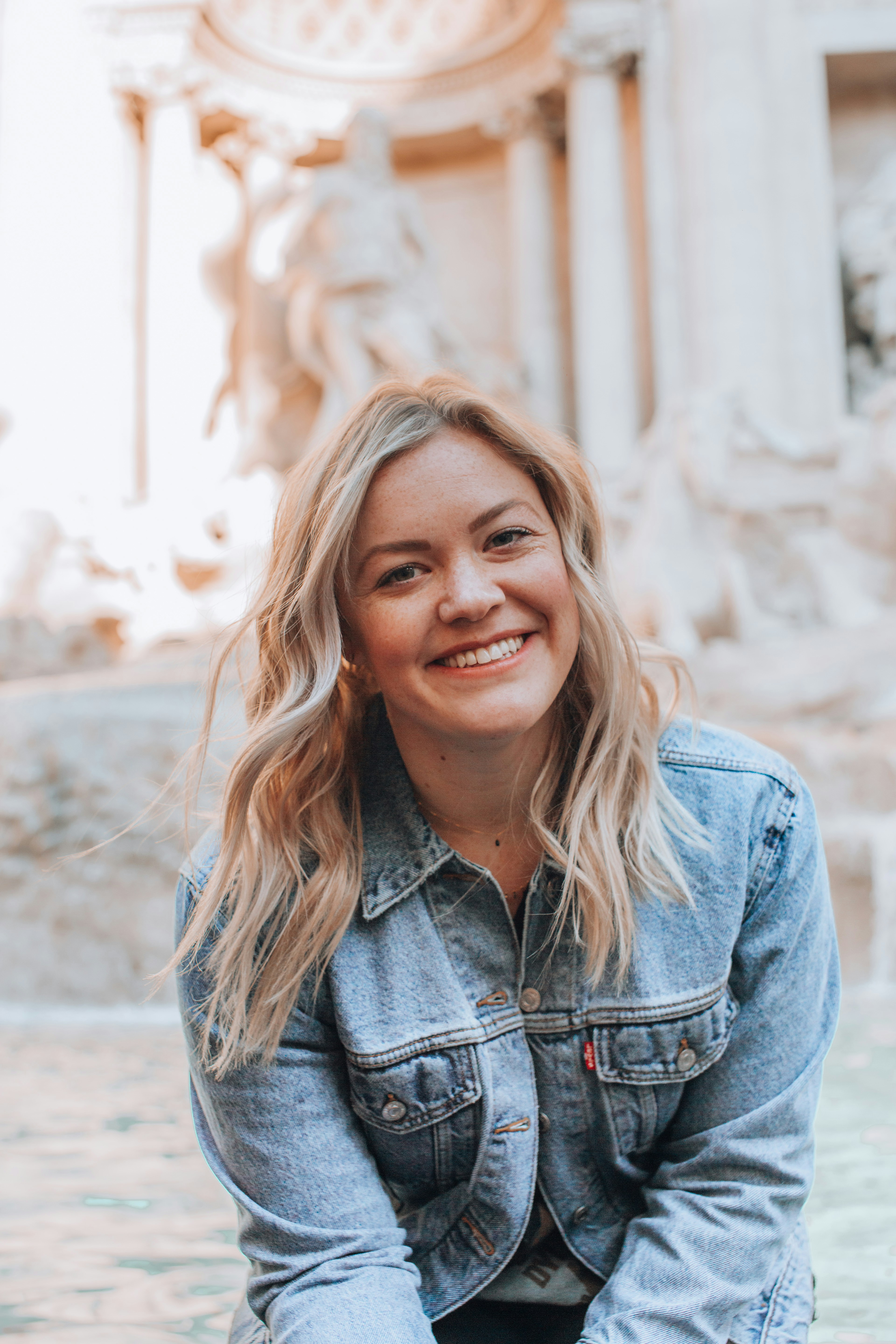 A smiling woman with long, wavy blonde hair, wearing a denim jacket, sitting in front of a historic fountain.